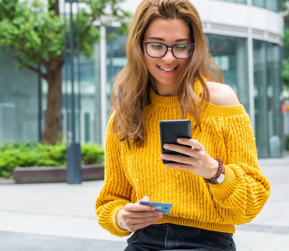 A young woman in a warm yellow sweater is smiling while typing their card number into their smart phone.